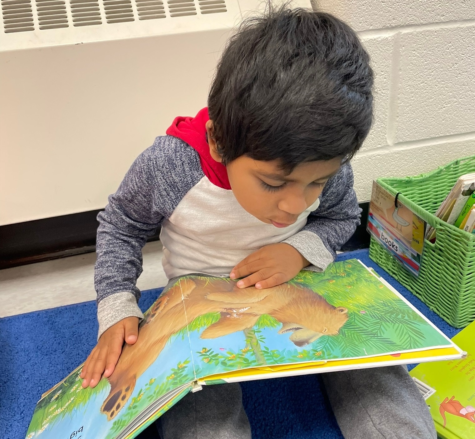 A toddler boy in a PLASP program sits on a blue carpet and looks at a picture book