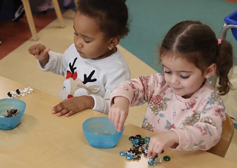 Two young children sit together at a table in a classroom and play with spare parts 