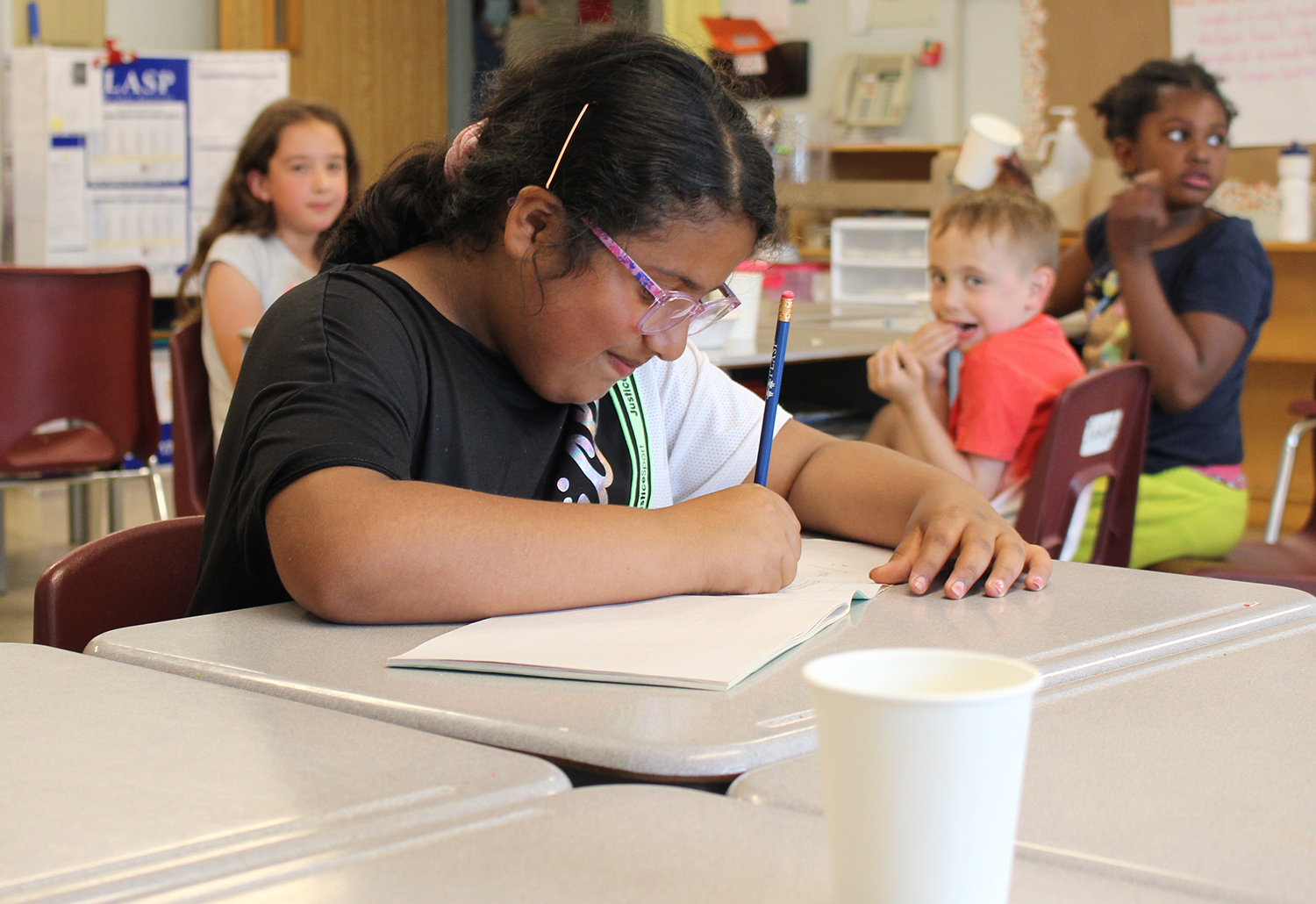 A girl in a PLASP school age after school program sits at a desk and writes in a notebook in a school classroom