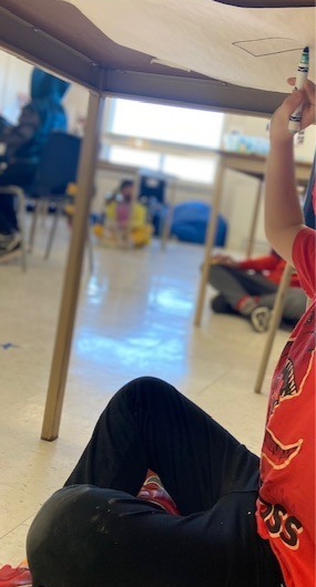 A child in a PLASP program sits under a desk that has a white piece of paper taped to it, and draws on it with a blue marker